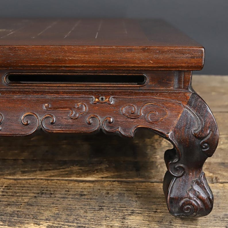 May include: A dark brown wooden display stand featuring a rectangular top surface and ornate, carved cabriole legs. The apron of the stand is decorated with intricate, swirling relief patterns and includes a horizontal rectangular cutout.