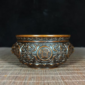 May include: A dark brown, round bronze incense burner featuring intricate relief carvings of swirling clouds, floral motifs, and circular medallions. The central medallion displays the Chinese character for treasure, surrounded by detailed scrollwork.