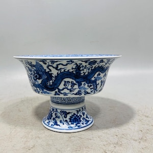 Op de afbeelding: Een witte porseleinen schaal op voet met gedetailleerde, met de hand geschilderde blauwe drakenmotieven onder het glazuur. De uitlopende rand en de voet zijn versierd met geometrische patronen, terwijl de steel florale ontwerpen toont.