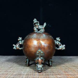 May include: A round bronze incense burner featuring a lid topped with a sculpted guardian lion figure. The vessel has two side handles shaped like mythical creatures and stands on three legs with ornate detailing and a geometric band pattern.
