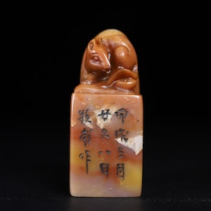 May include: A rectangular stone seal featuring a carved rat figure on top. The stone displays a mottled color palette of tan, brown, and cream. The front face of the seal is engraved with black Chinese characters arranged in three vertical columns.