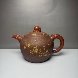 May include: A dark brown textured clay teapot featuring a gold-painted plum blossom branch design and the Chinese character for good fortune. The teapot has a dragon-shaped spout, a curved handle, and a lid topped with a small round knob.