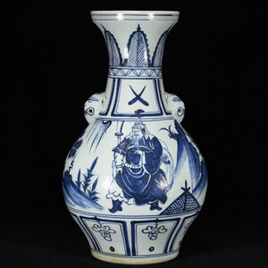 May include: A white porcelain vase featuring blue hand-painted illustrations of a standing figure in traditional attire, surrounded by landscape elements. The vase has two side handles and a flared rim, with decorative patterns at the neck and base.