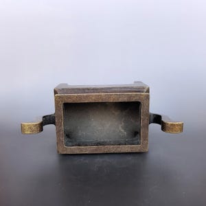 Pode incluir: Um queimador de incenso retangular de bronze com um acabamento met&aacute;lico escuro e desgastado. O recipiente apresenta um topo aberto e duas al&ccedil;as sim&eacute;tricas curvas que se estendem pelos lados, exibindo uma p&aacute;tina envelhecida e texturizada.
