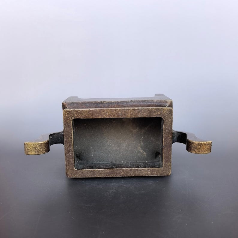 Pode incluir: Um queimador de incenso retangular de bronze com um acabamento met&aacute;lico escuro e desgastado. O recipiente apresenta um topo aberto e duas al&ccedil;as sim&eacute;tricas curvas que se estendem pelos lados, exibindo uma p&aacute;tina envelhecida e texturizada.