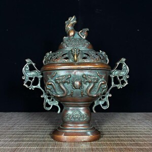May include: A bronze-toned metal incense burner featuring a lid topped with a mythical creature figure. The body displays embossed dragon motifs and a central sphere, flanked by two ornate, openwork handles on a pedestal base.
