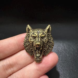 Puede incluir: Un broche de metal color latón con forma de cabeza de lobo gruñendo. El lobo se muestra con la boca abierta, revelando dientes afilados y un patrón de pelaje texturizado. El acabado metálico tiene áreas oscuras para resaltar los detalles faciales.