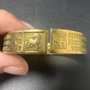 Puede incluir: Una pulsera tipo brazalete de metal dorado con una figura de caballo en relieve dentro de un marco cuadrado, flanqueada por secciones de caracteres chinos grabados y un sello cuadrado. La pulsera tiene un diseño rígido y abierto.