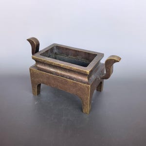 Pode incluir: Um queimador de incenso retangular de bronze com uma bacia profunda, uma borda larga e quatro pernas robustas. Duas al&ccedil;as curvas voltadas para cima estendem-se dos lados mais curtos do recipiente, que tem um acabamento met&aacute;lico escuro.