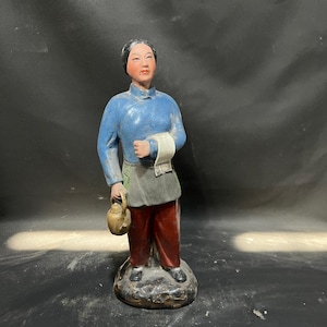 May include: A ceramic figurine of a person with black hair, wearing a blue long-sleeved top, a dark grey apron, and reddish-brown trousers. The figure holds a small tan teapot in one hand and a white cloth in the other, standing on a base.