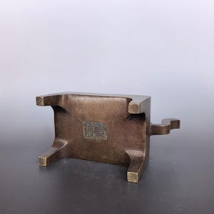 Pode incluir: Um suporte de metal em tom de bronze com quatro pernas curtas e uma base retangular. A parte inferior apresenta uma reentr&acirc;ncia quadrada central contendo um selo em relevo com marcas complexas. Uma pequena al&ccedil;a curva projeta-se de um lado.