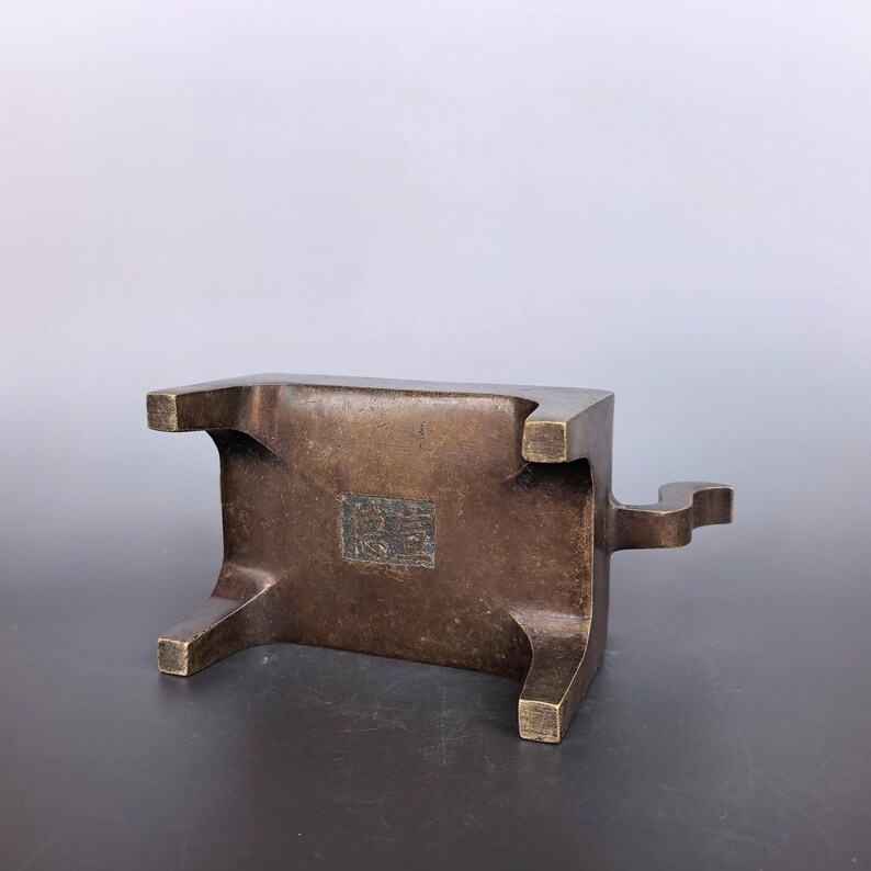 Pode incluir: Um suporte de metal em tom de bronze com quatro pernas curtas e uma base retangular. A parte inferior apresenta uma reentr&acirc;ncia quadrada central contendo um selo em relevo com marcas complexas. Uma pequena al&ccedil;a curva projeta-se de um lado.