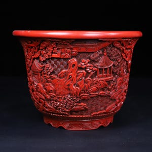 Puede incluir: Un cuenco de laca de cinabrio de color rojo intenso con intrincadas tallas en relieve de una escena paisajística. La superficie muestra representaciones detalladas de estructuras tradicionales, árboles estilizados, formaciones rocosas y patrones de agua.
