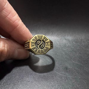 May include: A gold-toned metal ring featuring an octagonal face with a central yin-yang symbol surrounded by a series of trigram lines. The band of the ring displays intricate geometric patterns along the sides, providing a textured appearance.
