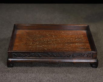 May include: A rectangular dark brown wooden display tray featuring a flat surface with a visible wood grain pattern. The tray has raised edges and a carved decorative border along the front side, supported by four short, curved feet at the corners.