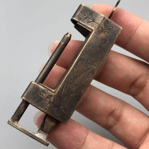 May include: Antique bronze-colored padlock with floral and bird engravings. The lock features a rectangular body with a sliding bar and a keyhole. The lock is held in a hand against a neutral background.