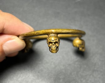 May include: A brass-toned metal cuff bracelet featuring three small, detailed skull charms attached to the outer band. The metal has a weathered, antique finish with visible texture on the skulls and the circular band structure.