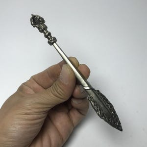 May include: A silver letter opener with ornate detailing. The handle features a decorative finial, and the blade has a pointed end with a detailed design. The opener is held in a hand against a white background.