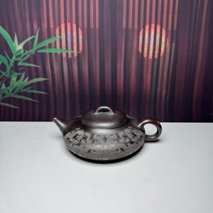 May include: A dark brown clay teapot featuring a low, rounded body and a matching lid with a small handle. The exterior surface is decorated with intricate, light-colored engraved patterns that wrap around the lower section of the teapot.
