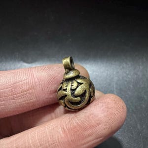 Puede incluir: Un peque&ntilde;o colgante esf&eacute;rico de lat&oacute;n con un intrincado dise&ntilde;o calado en relieve de patrones org&aacute;nicos y sinuosos. El metal tiene un acabado antiguo desgastado con una p&aacute;tina oscura en las &aacute;reas hundidas y una peque&ntilde;a anilla superior.