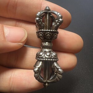 Puede incluir: Un colgante vajra de metal plateado con intrincadas tallas simétricas, una sección de agarre central y extremos dentados que se curvan hacia adentro para encontrarse en una punta, con un acabado pulido y detalles oscuros.