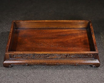 May include: A rectangular dark brown wooden tray with a smooth, polished interior surface and raised edges. The front exterior side features intricate, low-relief carvings of floral patterns and branches, supported by four small, curved feet.