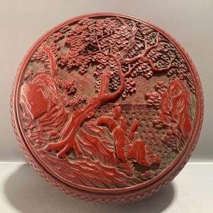 May include: A round red cinnabar lacquer box featuring an intricate relief carving on the lid. The design depicts a detailed landscape with a large tree, jagged rock formations, and a figure seated in a garden setting with a patterned fence background.