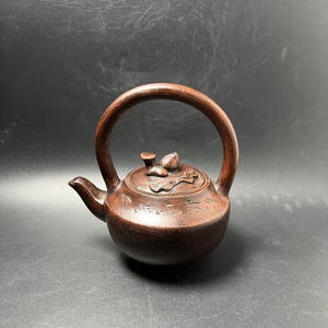 May include: A dark brown, textured clay teapot featuring a high, arched handle and a short spout. The lid is adorned with a sculpted leaf and bud motif, while the body of the teapot displays engraved Chinese characters around the upper section.