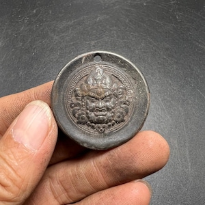 May include: A circular dark gray metal pendant featuring a raised relief of a fierce deity face at the center, surrounded by a decorative border pattern and a small hole at the top for suspension. The surface has a weathered, matte finish.