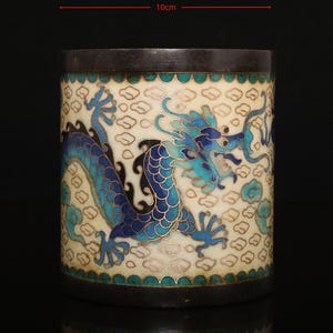 May include: A cylindrical cloisonné brush pot featuring a blue and turquoise dragon motif against a cream-colored background with cloud patterns. The vessel has dark metal rims and measures 10 cm in diameter and 11 cm in height.