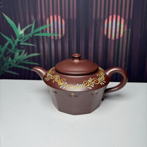 Puede incluir: Una tetera de arcilla marrón oscuro con base hexagonal y tapa a juego. La parte superior presenta un patrón decorativo pintado de enredaderas amarillas y verde claro, con pequeños puntos amarillos que acentúan el borde inferior del diseño.