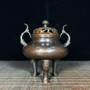 May include: A bronze-toned metal incense burner featuring a rounded body, three sturdy legs with engraved detailing, and two curved side handles. The piece includes a removable lid with an intricate openwork design and a small central knob.