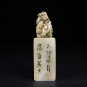 May include: A rectangular off-white stone seal featuring a carved mythical creature on top. The front face of the seal displays vertical columns of engraved Chinese characters. The stone has a smooth, polished texture with subtle greyish marbling throughout the surface.