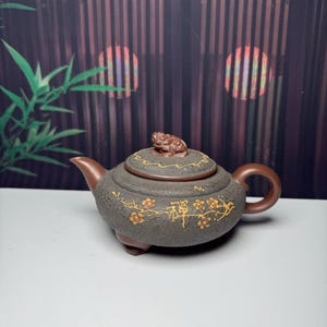 May include: A dark grey textured teapot with a smooth brown handle and spout. The lid features a small sculpted animal figure. The body is decorated with gold-coloured painted plum blossoms and a central Chinese character. It stands on three feet.