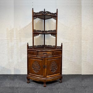 May include: A dark brown wooden corner cabinet featuring two open upper shelves with intricate carved lattice detailing. The base includes a central drawer with a carved front and two lower cabinet doors, each displaying a circular carved motif.