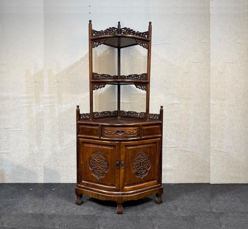 May include: A dark brown wooden corner cabinet featuring two open upper shelves with intricate carved lattice detailing. The base includes a central drawer with a carved front and two lower cabinet doors, each displaying a circular carved motif.