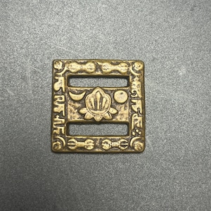 May include: A square brass-toned metal buckle featuring a central embossed lotus flower motif flanked by a crescent moon and a circle. The frame is decorated with repeating geometric patterns and small circular accents along all four edges.