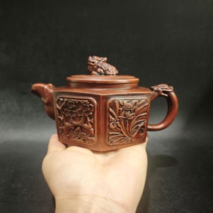 Peut inclure: Une théière hexagonale en argile brune ornée de sculptures en relief représentant des motifs floraux sur ses côtés. Le couvercle est surmonté d'une petite créature sculptée, tandis que l'anse et le bec présentent des motifs animaliers détaillés.