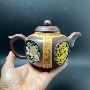 May include: A brown clay teapot featuring a central yellow band with intricate scrollwork patterns. The body includes two circular cutouts, one displaying a green turtle relief and the other a yellow dragon relief, topped with a matching lid.