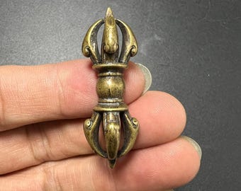 May include: A small, antique-style brass vajra pendant featuring a central spherical bead flanked by symmetrical, curved prongs that meet at pointed tips on both ends. The metal has a weathered, darkened patina with visible highlights.