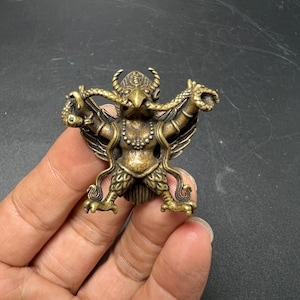 May include: A small, antique-style brass figurine depicting a mythical creature with a bird-like head, wings, and horns. The figure is shown holding a long, slender serpent in its hands, with the snake's body curving around the creature's arms.