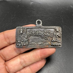 May include: A rectangular dark gray metal pendant featuring a central loop at the top. The surface displays embossed figures of two seated deities on the left and right sides, with Sanskrit script characters centered between them.