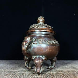 K&ouml;nnte beinhalten: Ein bronzefarbener R&auml;uchergef&auml;&szlig; aus Metall mit einem bauchigen K&ouml;rper, drei Beinen in Elefantenkopf-Form und einem passenden Deckel. Der Deckel ist mit einem filigranen, durchbrochenen Blumenmuster versehen und der obere Rand ist mit Ranken verziert.