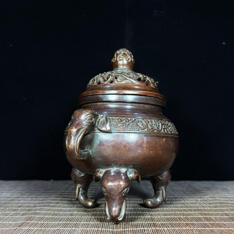 K&ouml;nnte beinhalten: Ein bronzefarbener R&auml;uchergef&auml;&szlig; aus Metall mit einem bauchigen K&ouml;rper, drei Beinen in Elefantenkopf-Form und einem passenden Deckel. Der Deckel ist mit einem filigranen, durchbrochenen Blumenmuster versehen und der obere Rand ist mit Ranken verziert.