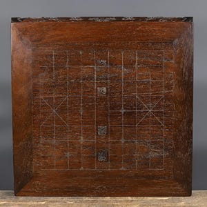 May include: A square dark brown wooden board featuring an inlaid grid pattern for Chinese chess. The surface displays a natural wood grain texture with thin, light-colored lines forming the game board layout and Chinese characters in the center.