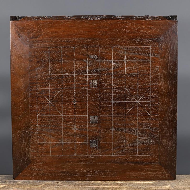 May include: A square dark brown wooden board featuring an inlaid grid pattern for Chinese chess. The surface displays a natural wood grain texture with thin, light-colored lines forming the game board layout and Chinese characters in the center.