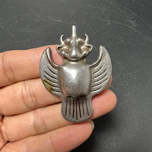 Pode incluir: Um broche de metal prateado em forma de escaravelho alado estilizado. A peça apresenta um corpo central arredondado, asas simétricas com detalhes de linhas gravadas e uma seção de cabeça proeminente com duas pequenas protuberâncias em forma de chifre.