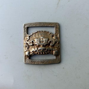 May include: A silver-colored rectangular buckle with an ornate, detailed design of a mythical creature's face. The creature has a fierce expression with visible teeth and intricate swirling patterns. The buckle's frame has a textured border.