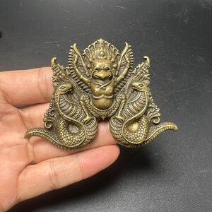 Pode incluir: Um ornamento de metal em tom de latão com uma figura central alada com rosto de pássaro, ladeada por duas criaturas simétricas em forma de serpente com escamas texturizadas. A peça apresenta detalhes intrincados em relevo.