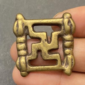 May include: A square, antique brass pendant featuring a stylized swastika design. The pendant has a detailed, openwork pattern with rounded edges and decorative elements at each corner. The brass has a warm, aged patina.